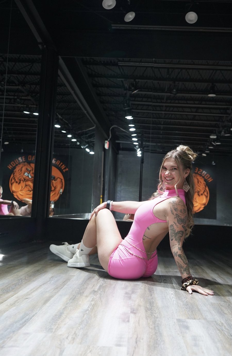 Sami in a pink workout set, sitting on the studio floor and smiling over her shoulder